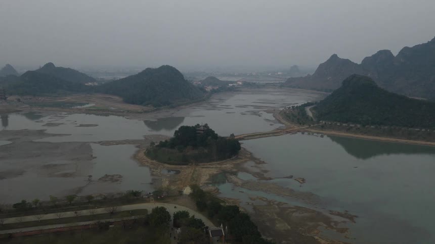 An atmospheric aerial view slowly approaches an ancient pagoda on a small island in a vast, misty lake surrounded by majestic mountains