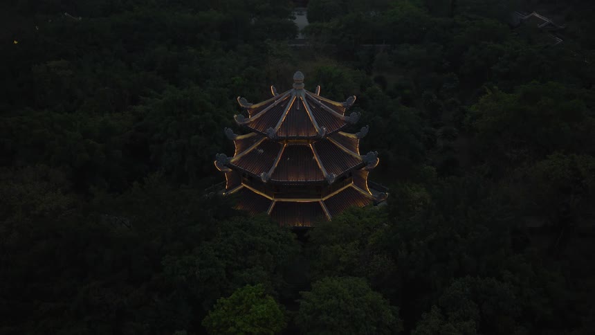 A striking aerial top-down view slowly zooms in on a beautifully illuminated ancient pagoda nestled in a dense, dark forest at twilight