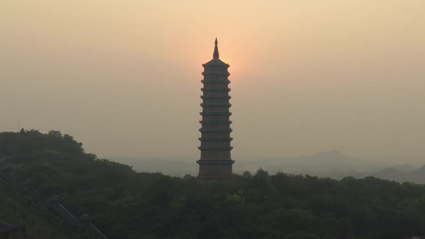 A time-lapse captures the sun rising from behind a tall Buddhist pagoda during a hazy, golden sunrise