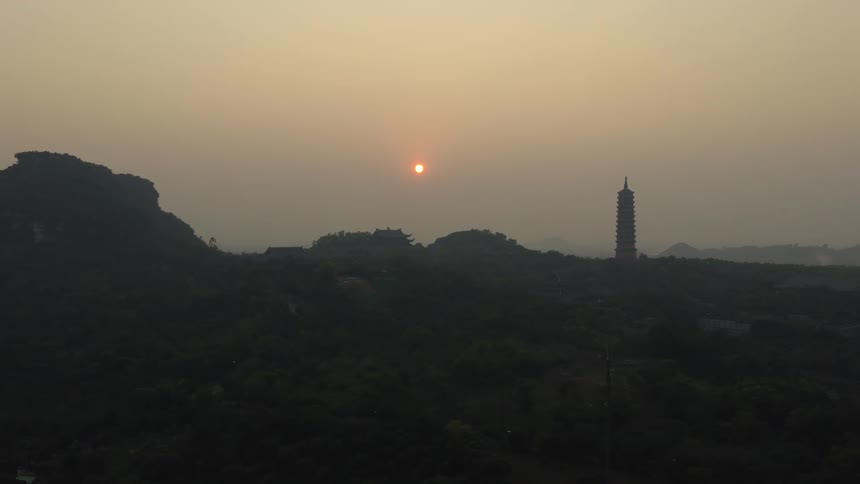 A hazy sunset glows over a vast green landscape featuring a traditional Asian pagoda