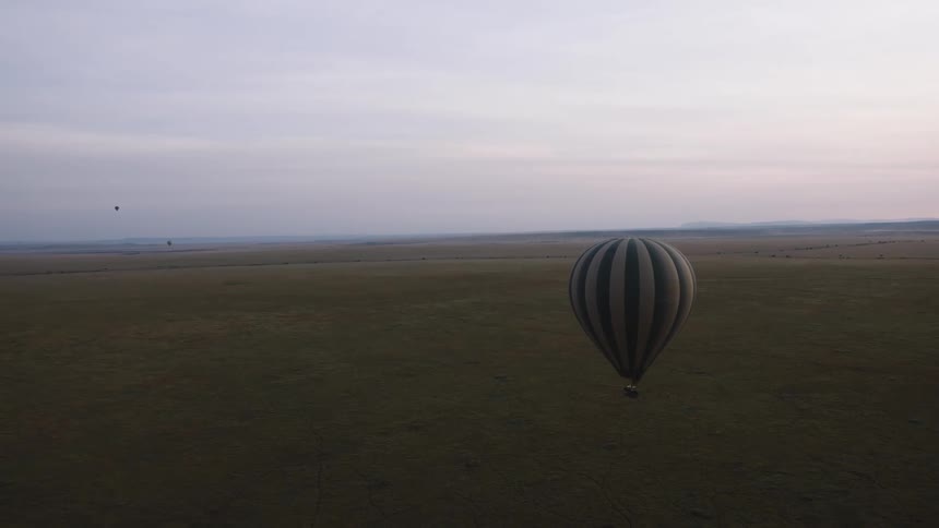 A hot air balloon with black and white stripes floats over the vast plains of the savanna during a hazy sunrise