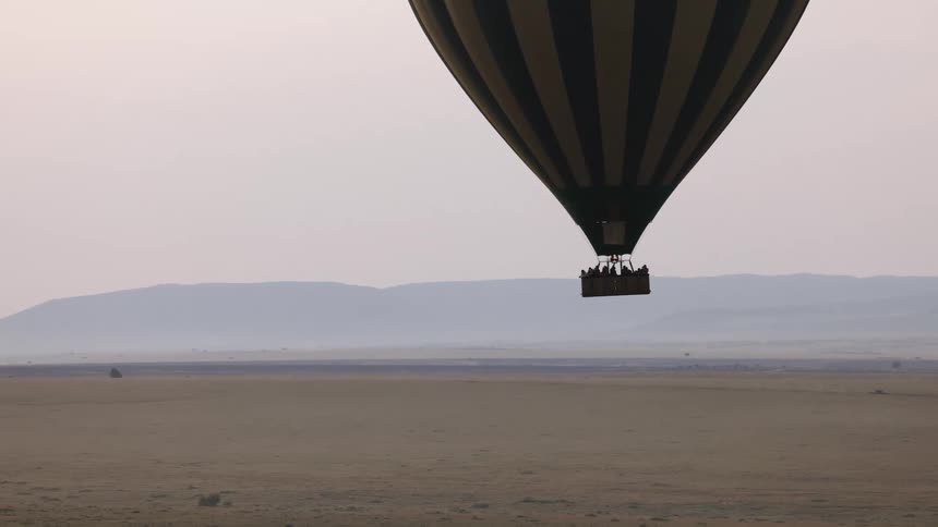 A striped hot air balloon full of tourists glides gracefully over the hazy African savanna with mountains in the distance