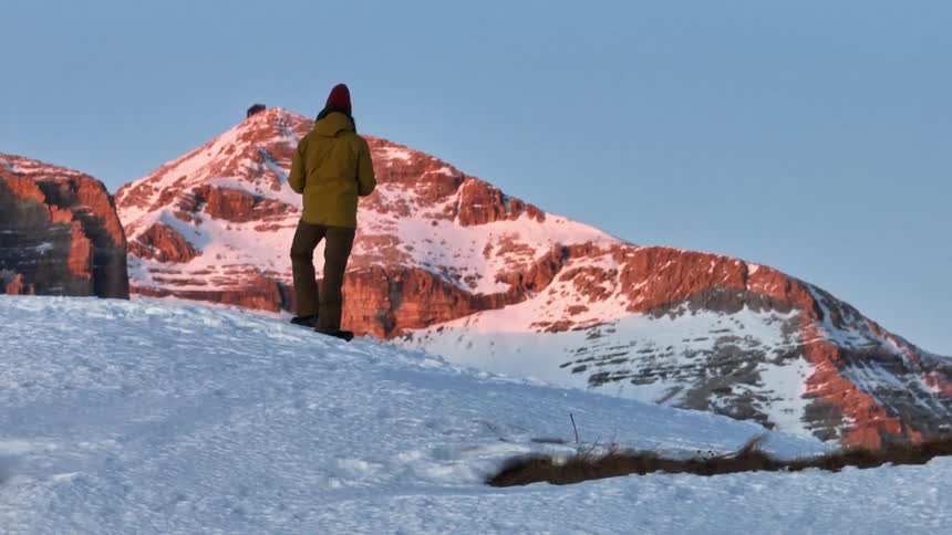 A drone pilot on a snowy ridge launches a drone, revealing a breathtaking vista of the snow-covered Dolomite mountains illuminated by the golden sunrise