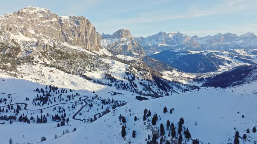 A breathtaking aerial view reveals a vast, sunlit valley and winding mountain pass in the snow-covered Alps