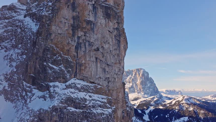 A dramatic aerial shot pulls back from a massive rock cliff to reveal the breathtaking, snow-covered Dolomite mountains