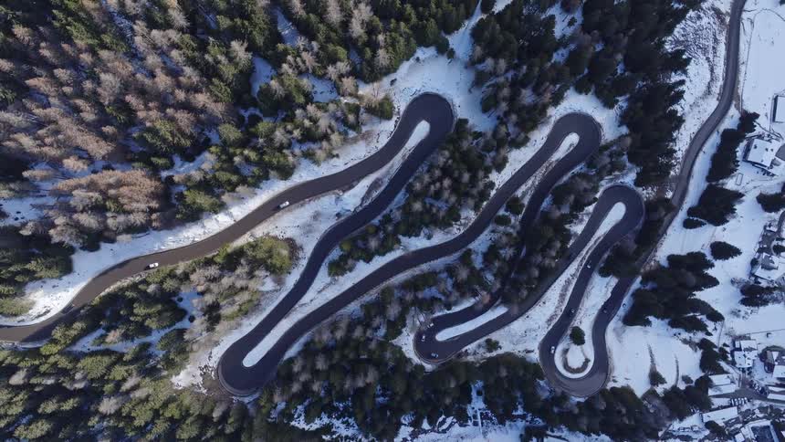 An aerial top-down view shows cars driving along a winding mountain pass road with many switchbacks through a snowy forest