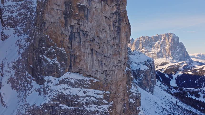 Epic aerial footage pulling back from a sheer, snow-dusted mountain cliff to reveal the majestic alpine landscape on a clear winter day
