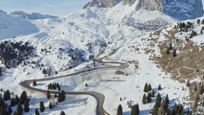 A stunning aerial view follows cars driving on a winding mountain road, tilting up to reveal the majestic snow-covered peaks of the Dolomites