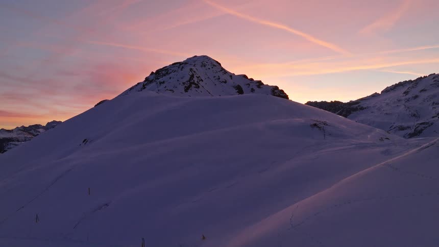A camera tilts up a pristine, snow-covered mountain slope to reveal a majestic peak against a beautiful pink and purple sunset sky