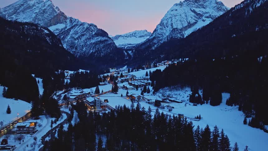 An aerial view of a tranquil, snow-covered village in a mountain valley at twilight, with a slow tilt up revealing the majestic peaks against a colorful sky