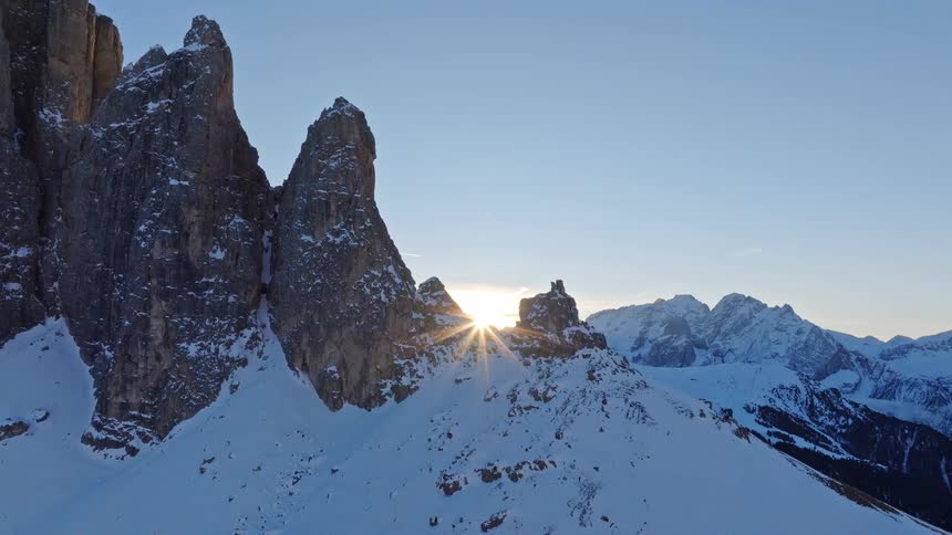 A stunning aerial drone shot pulls back to reveal a brilliant sunrise peeking from behind the jagged, snow-covered peaks of a majestic mountain range