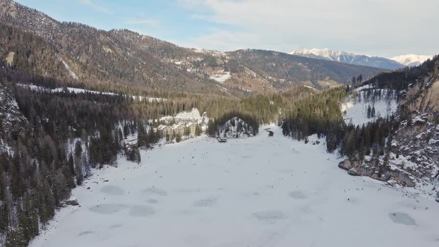 A stunning pull-back aerial shot reveals a vast frozen lake where people are walking and playing, surrounded by snowy mountains and a dense pine forest