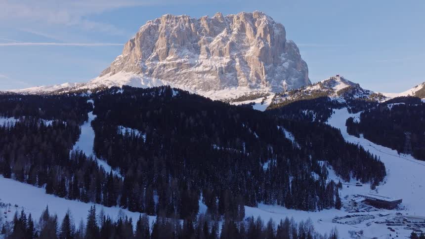 An aerial view slowly reveals a majestic sun-kissed mountain peak above a snow-covered forest and ski slopes