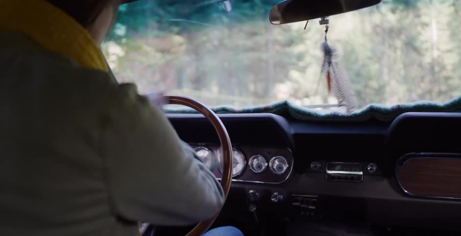 Over the shoulder view of a woman driving a classic car down a scenic forest road