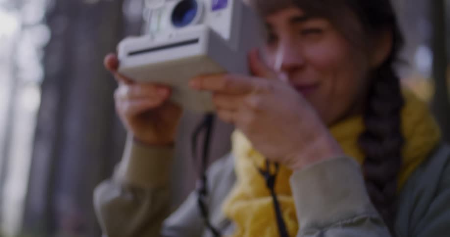 A young woman with a yellow scarf smiles and winks as she lifts a Polaroid instant camera to take a photo in the woods