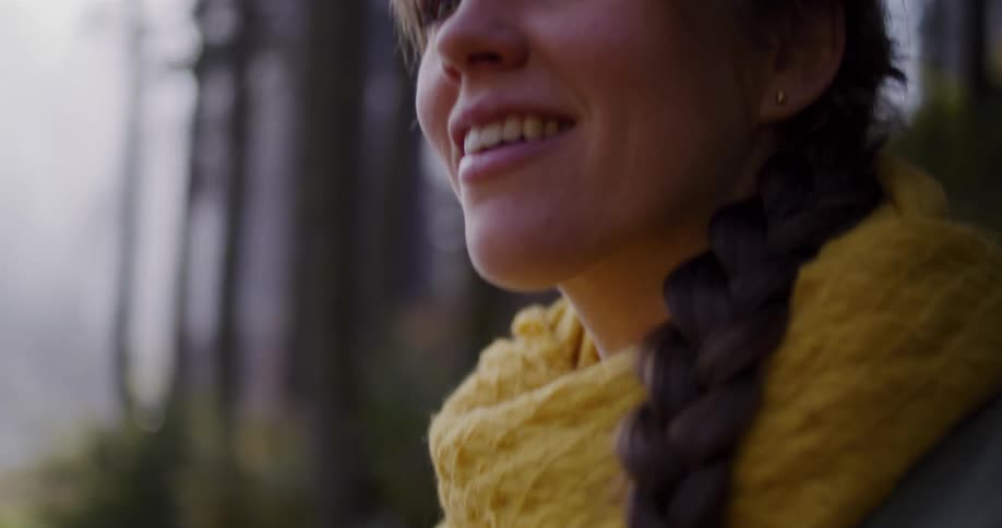 A serene young woman with a long braid and a yellow scarf smiles while enjoying the peaceful forest