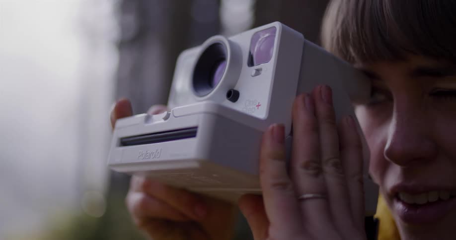 A young woman with green eyes smiles as she raises a Polaroid camera to take a picture while standing in a forest