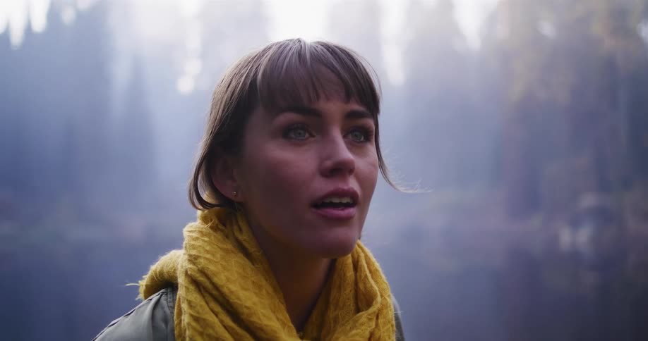 A beautiful young woman wearing a yellow scarf looks up in amazement while standing in a foggy, atmospheric forest