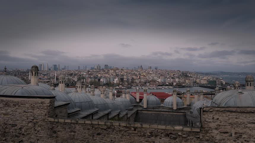 A wide, static view from a historic rooftop with grey domes overlooks the modern skyline of Istanbul, Turkey, under a moody, overcast sky