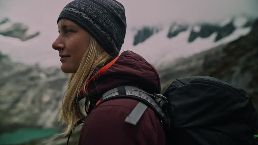 A contemplative female hiker with a backpack pauses to admire the magnificent view of a snow-capped mountain range in slow motion