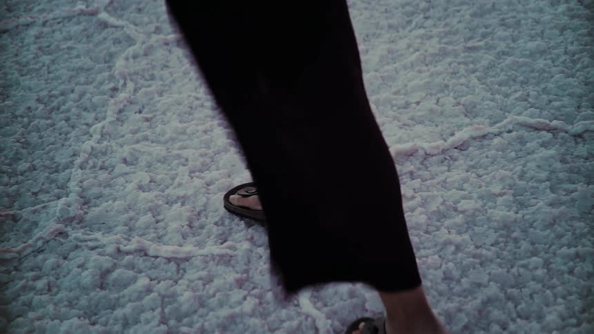 A slow motion, high-angle view of a person's feet in sandals walking across a pink salt flat