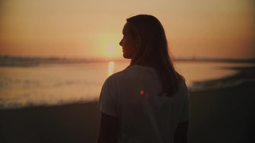 A woman in silhouette stands peacefully on the beach, looking out at the beautiful golden sunset over the ocean