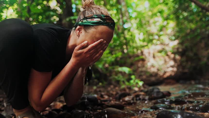 A woman crouches by a forest creek, scooping up clear water with her hands and washing her face in slow motion