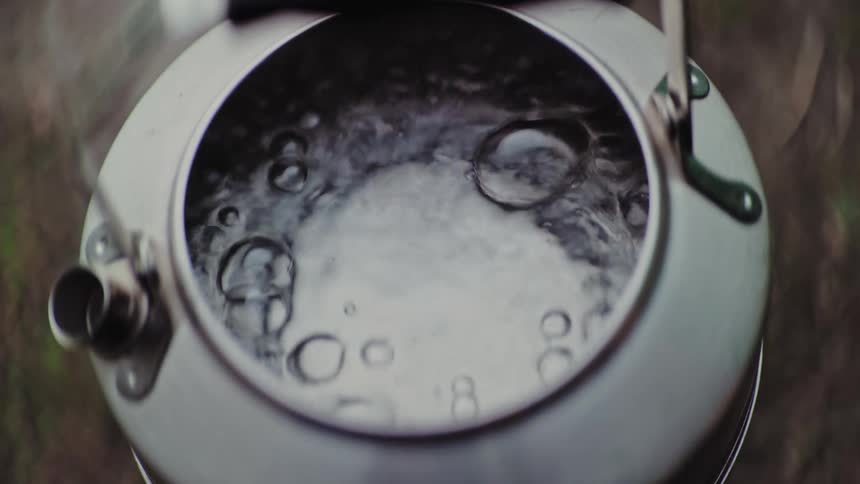 A close-up overhead shot shows water bubbling and boiling in a silver camping kettle in slow motion