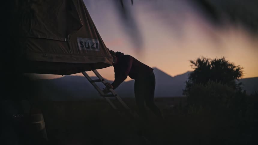 A female traveler climbs down from her rooftop tent, silhouetted against a stunning mountain sunrise