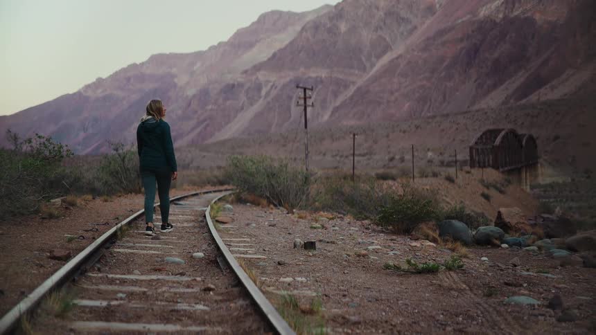 A solitary woman walks down abandoned railway tracks through a scenic mountain valley at dusk