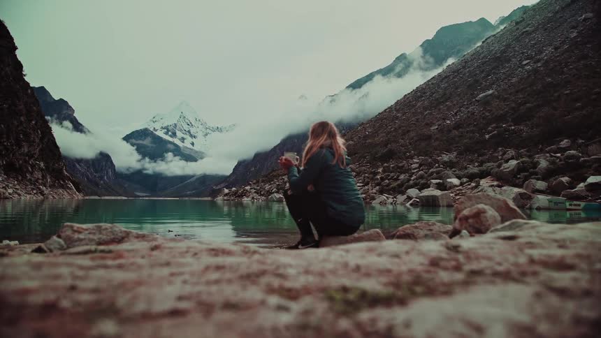 A woman in a jacket squats by a tranquil, turquoise lake in the mountains, holding a mug and enjoying the scenic view