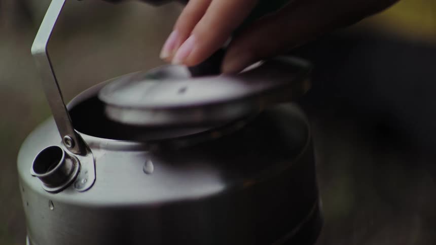 Slow motion close-up of a hand carefully placing the lid on a steaming metal camping kettle