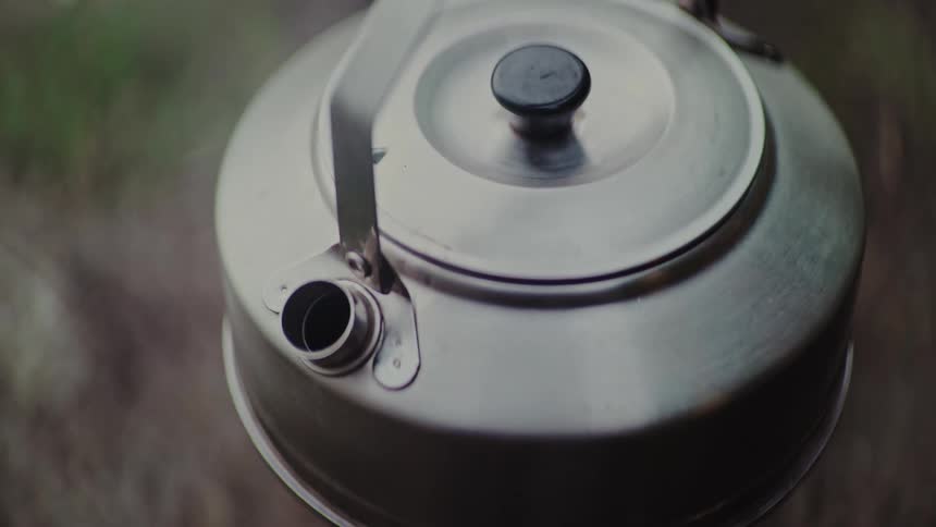 A top-down close-up shot of a stainless steel camping kettle boiling water, with steam rising from the spout