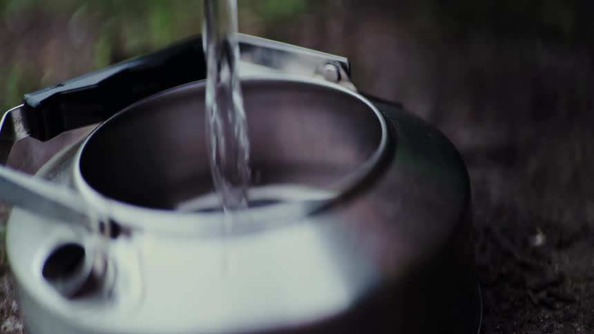 A slow-motion close-up of water being poured into a steaming stainless steel camping kettle outdoors
