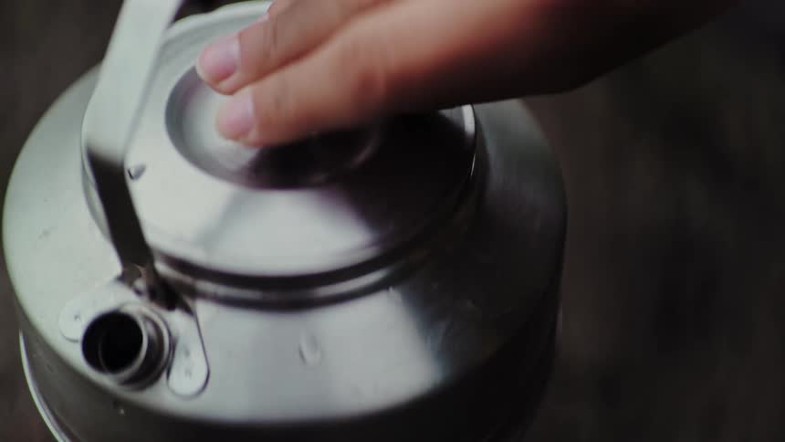 A person's hand carefully places the lid on a steaming stainless steel camping kettle as water boils inside