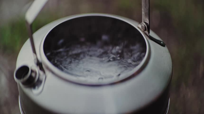 A hand removes the lid of a metal camping kettle in slow motion, revealing the vigorously boiling water and steam inside