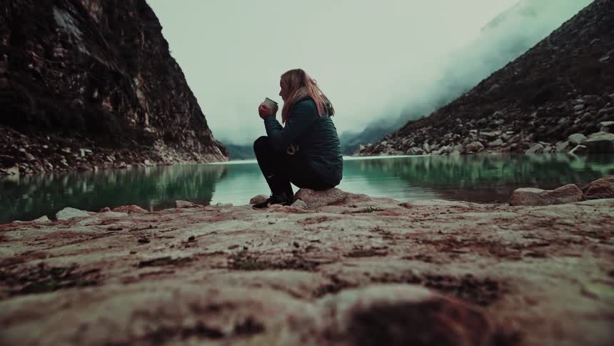 A woman contemplates the view while sipping a warm drink by a tranquil, turquoise mountain lake on a foggy day