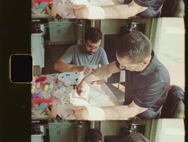 Vintage 8mm style split-screen footage of two men enjoying a creative painting session together at a sunlit kitchen table
