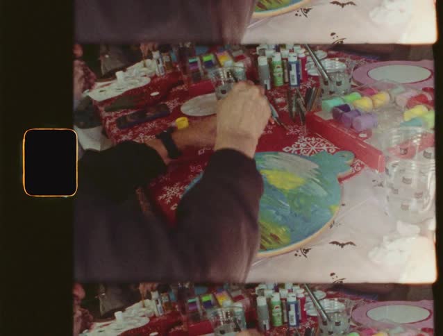 Vintage 8mm footage captures hands painting a colorful, large ornament at a table filled with holiday craft supplies