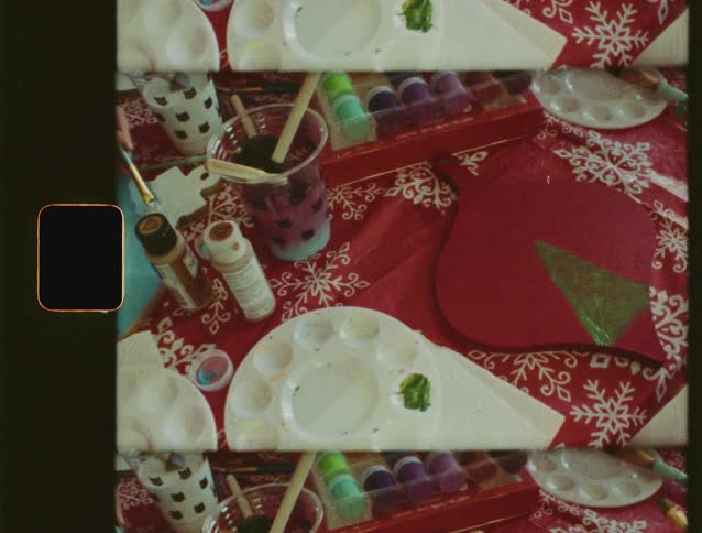 A vintage, split-screen view from above shows hands painting Christmas decorations on a festive tablecloth