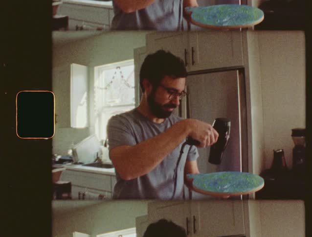 A man smiles for the camera then uses a hair dryer to dry his planet Earth art project in a vintage film-style split-screen video