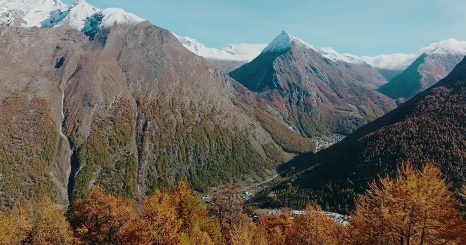 An aerial shot flies over a golden autumn forest to reveal a picturesque alpine village nestled in a valley surrounded by snow-capped mountains