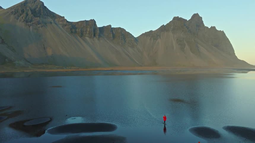 A stunning aerial view of a person in a red jacket standing in a tranquil tidal lagoon, with the majestic Vestrahorn mountain reflecting in the water in Iceland