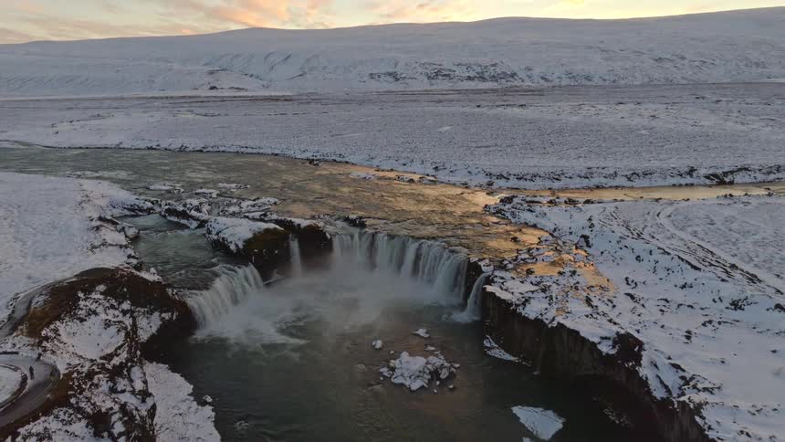 A majestic aerial view reveals the powerful Goðafoss waterfall cascading through a snowy Icelandic landscape at sunset