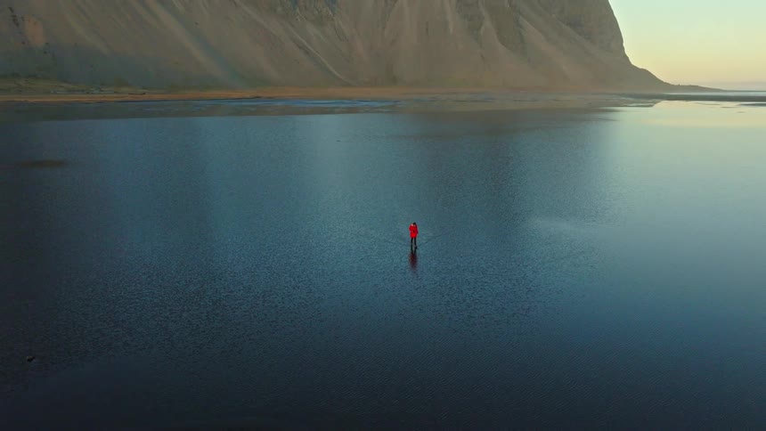 A stunning aerial shot reveals a person in a red jacket standing alone in a vast, calm body of water, reflecting the epic Vestrahorn mountain in Iceland at sunrise
