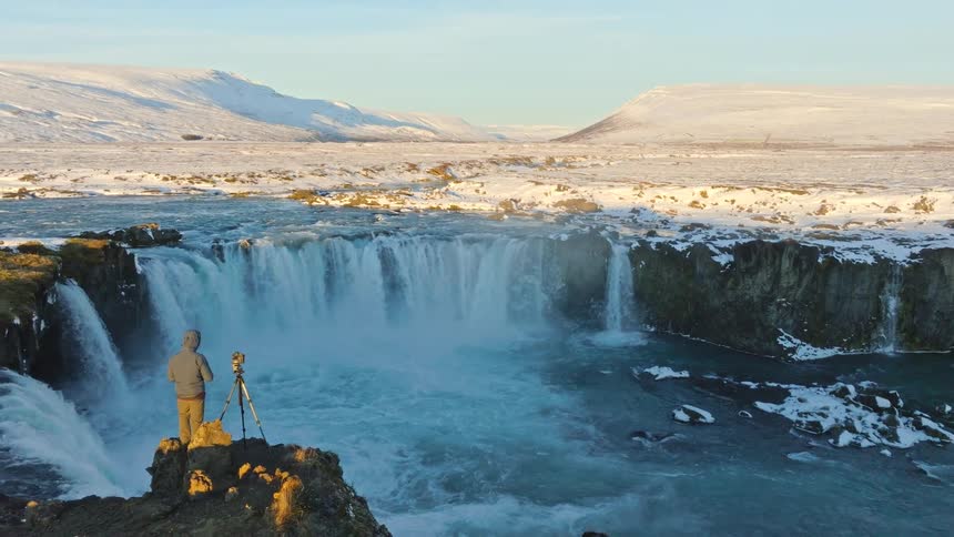 A lone photographer stands on a cliff's edge capturing the majestic Goðafoss waterfall in a vast, snowy Icelandic landscape at sunrise