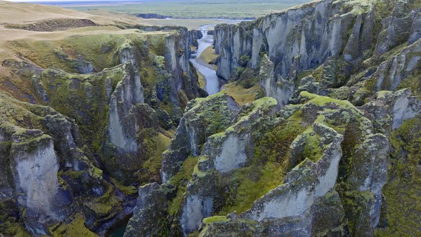 A slow aerial pullback reveals the epic scale of a deep, mossy canyon with a winding river flowing through its base in the Icelandic highlands