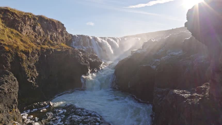 An aerial drone shot slowly rises to reveal a powerful waterfall flowing through a rugged canyon with the sun creating a misty haze