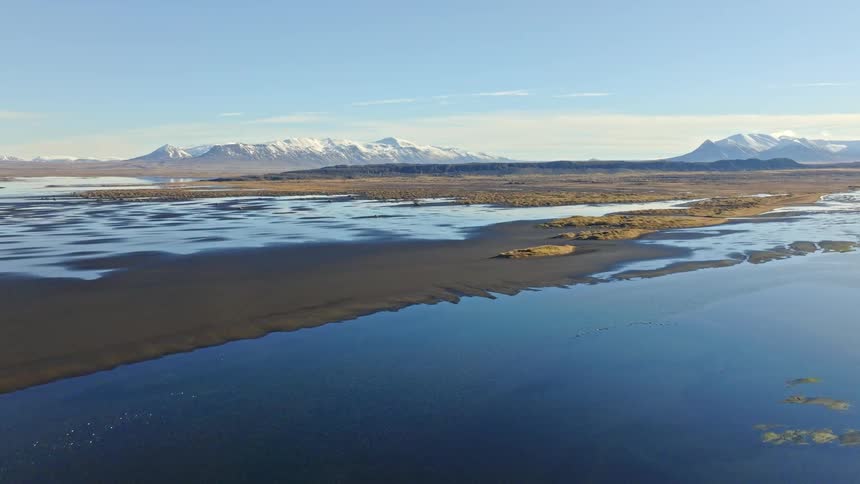 Stunning aerial ascent over a tranquil Icelandic lake, revealing a black sand shoreline and majestic snow-capped mountains under a clear blue sky
