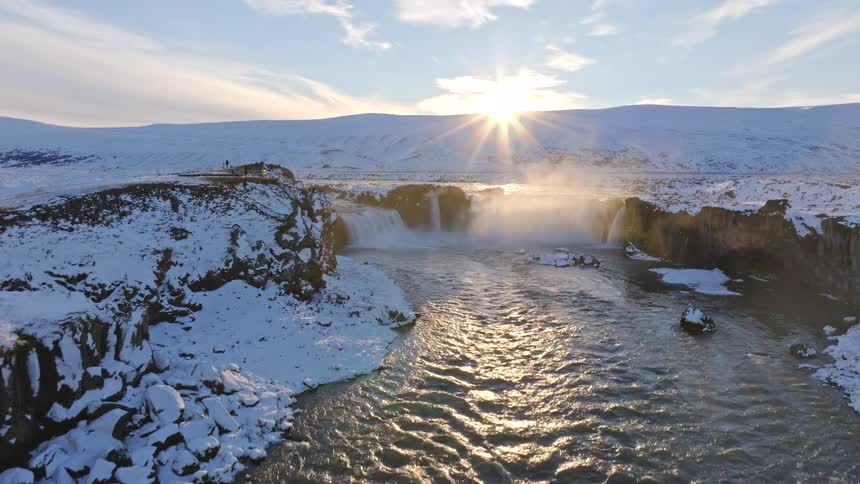 An aerial drone shot rises over a powerful river to reveal the majestic Goðafoss waterfall in a snowy Icelandic landscape at sunset
